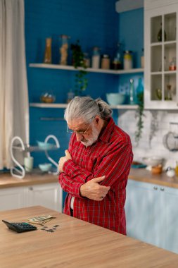 Aydınlık bir mutfakta masanın üstünde kırmızı gömlekli yaşlı bir adam masraflarını aylık fatura olarak sayar.