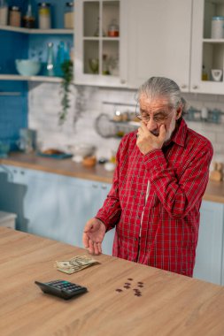 Aydınlık bir mutfakta masanın üstünde kırmızı gömlekli yaşlı bir adam masraflarını aylık fatura olarak sayar.