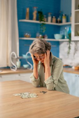 Parlak bir mutfakta açık yeşil tişörtlü yaşlı bir kadın para dolu bir masanın yanında duruyor.