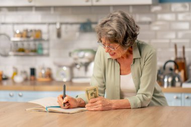 Parlak bir mutfakta açık yeşil tişörtlü yaşlı bir kadın, içinde para olan bir masanın yanında duruyor. Birikimleri sayıyor.