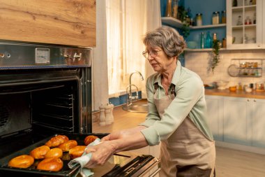 Parlak bir mutfakta açık yeşil tişörtlü yaşlı bir kadın fırından taze turtalar çıkarıyor. Mutfakta mutlu, yaşlı bir ev hanımı.