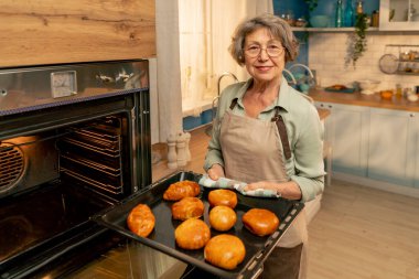 Parlak bir mutfakta açık yeşil tişörtlü yaşlı bir kadın fırından taze turtalar çıkarıyor. Mutfakta kameraya bakıyor.
