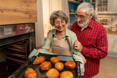 Parlak bir mutfakta açık yeşil tişörtlü yaşlı bir kadın fırından taze turtalar çıkarıyor mutfaktaki mutlu yaşlı ev kadını kocası arkadan sarılıyor.