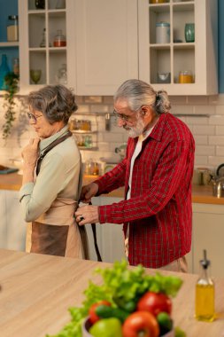 Parlak bir mutfakta kırmızı gömlekli yaşlı bir adam yemek yapmadan önce arkasından gülümsüyor. Karısı için önlük bağlıyor. Mutlu bir çift.