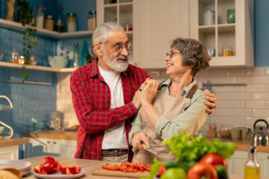 Parlak bir mutfakta, açık yeşil gömlekli yaşlı bir kadın kocasıyla sağlıklı bir yemek hazırlıyor, sebze kesiyor, koca da doğranmış biberi deniyor.