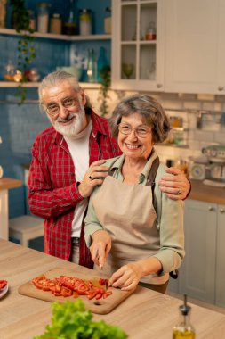 Parlak bir mutfakta açık yeşil gömlekli yaşlı bir kadın kocasıyla sağlıklı bir yemek hazırlıyor ve kameraya bakarak sebze kesiyor.
