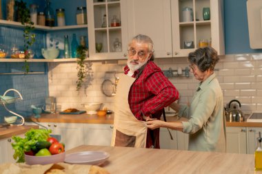 Parlak bir mutfakta yaşlı bir adam yeşil gömlekli yaşlı bir kadını pişirmeden önce kocasına bir önlük bağlıyor mutlu bir çift için.