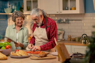 Parlak bir mutfakta, açık yeşil gömlekli yaşlı bir kadın kocasıyla sağlıklı bir akşam yemeği hazırlıyor koca sebze kesiyor kadın tabletten bir tarif izliyor.