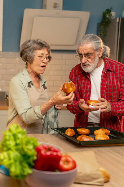 in a bright kitchen an elderly woman in a light green shirt is happy about the prepared pies her husband is happy tasting pastries