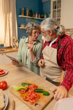 Parlak bir mutfakta, açık yeşil gömlekli yaşlı bir kadın kocasıyla sağlıklı bir akşam yemeği hazırlıyor koca sebze kesiyor kadın tabletten bir tarif izliyor.