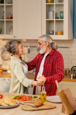Parlak bir mutfakta açık yeşil tişörtlü yaşlı bir kadın kocasıyla birlikte önlük giymiş mutfakta durmuş romantik bir şekilde gülümsüyor ve dans ediyor.