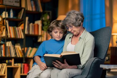 Oturma odasının sıcak ışığı. Büyük mavi koltuklu yaşlı kadın torunuyla oturuyor. Okumak için kitap okuyor.