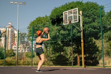 Kısa şortlu, siyah tişörtlü, kısa saçlı ve çeviklik konusunda basketbol antrenmanına basket topu atmak için spor sahasındaki aktif eğlence kızı.