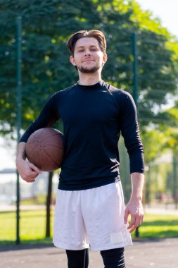 Spor sahası basketbol sahasında beyaz şortlu, siyah tişörtlü, elinde topla kameranın önünde poz veren adam.