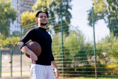 Spor sahası basketbol sahasında beyaz şortlu, siyah tişörtlü, elinde topla kameranın önünde poz veren adam.