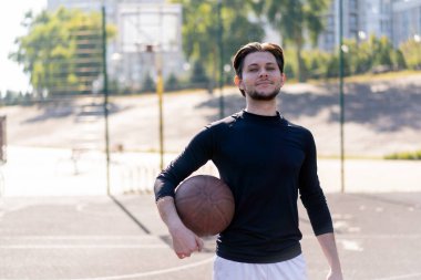 Spor sahası basketbol sahasında beyaz şortlu, siyah tişörtlü, elinde topla kameranın önünde poz veren adam.