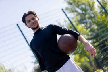 Spor sahası basketbol sahasında beyaz şortlu, siyah tişörtlü, elinde topla kameranın önünde poz veren adam.