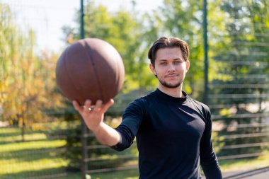 Spor sahasında, beyaz şortlu, siyah tişörtlü, elinde topla kameranın önünde poz veren aktif sporcu.