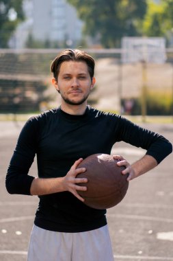 Spor sahası basketbol sahasında beyaz şortlu, siyah tişörtlü, elinde topla kameranın önünde poz veren adam.