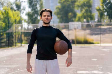 Spor sahası basketbol sahasında beyaz şortlu, siyah tişörtlü, elinde topla kameranın önünde poz veren adam.