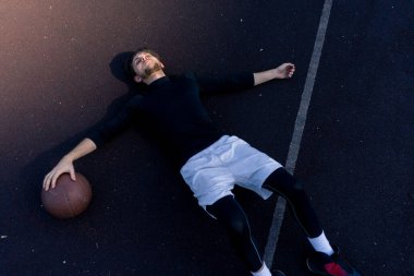 Spor sahası basketbol sahasında beyaz şortlu ve siyah tişörtlü adam topun yanındaki sahada yatıyor.