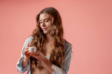 Stüdyo güzellik çekimi pembe arka planda uzun saçlı kız iç çamaşırı ve mavi gömlekli doğal güzellik kavanozuyla poz veriyor.