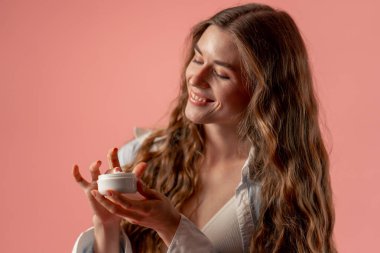 Stüdyonun güzellik çekimini kapat pembe arka planda uzun saçlı kız iç çamaşırı ve mavi gömlekli doğal güzellik kavanozuyla poz ver.