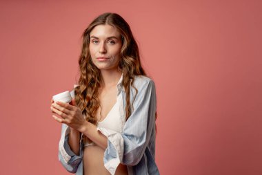 Stüdyo güzellik çekimi pembe arka planda uzun saçlı kız iç çamaşırı ve mavi gömlekli doğal güzellik kavanozuyla poz veriyor.