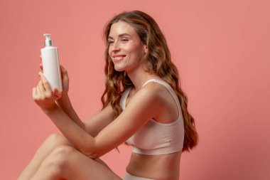 Stüdyonun güzellik çekimini kapat pembe arka planda uzun saçlı kız iç çamaşırıyla oturmuş diz kapağında losyonla poz veriyor doğal güzellik.