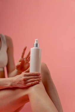 Stüdyonun güzellik çekimini kapat pembe arka planda uzun saçlı kız iç çamaşırıyla oturmuş, elinde losyon kutusuyla dizinde doğal güzellik var.