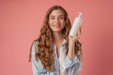 Stüdyo güzelliği, pembe arka planda uzun saçlı, mavi gömlekli, iç çamaşırlı, elinde losyon kutusuyla kameraya bakan doğal güzellik.