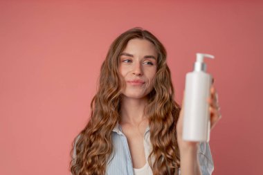 Stüdyo güzelliği, pembe arka planda uzun saçlı, mavi gömlekli, iç çamaşırlı, elinde losyon kutusuyla kameraya bakan doğal güzellik.
