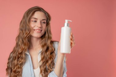 Stüdyo güzelliği, pembe arka planda uzun saçlı, mavi gömlekli, iç çamaşırlı, elinde losyon kutusuyla kameraya bakan doğal güzellik.