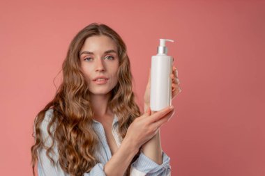 Stüdyo güzelliği, pembe arka planda uzun saçlı, mavi gömlekli, iç çamaşırlı, elinde losyon kutusuyla kameraya bakan doğal güzellik.