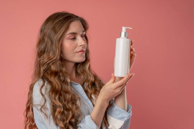 Stüdyo güzelliği, pembe arka planda uzun saçlı, mavi gömlekli, iç çamaşırlı, elinde losyon kutusuyla kameraya bakan doğal güzellik.