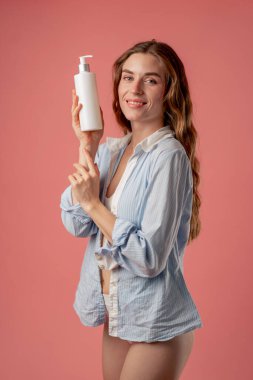 Stüdyonun güzellik çekimi pembe arka planda uzun saçlı kız mavi gömlekli, losyon kutusuyla poz veriyor doğal güzellik.