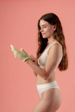 studio beauty shoot on a pink background beautiful girl in white underwear putting on a green massage glove for massaging her butt and thigh cellulite prevention beauty salon