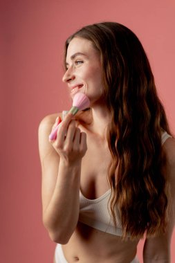 Stüdyo güzellik çekimi pembe arka planda beyaz iç çamaşırlı güzel bir kız pembe makyaj fırçalarıyla ayakta duruyor güzellik salonu gülümsüyor
