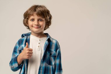 Beyaz arka planda bir fotoğraf stüdyosunda beyaz kazaklı ve mavi kareli gömlekli gülümseyen bir okul çocuğu kameraya bakar.