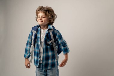 Beyaz arka planda bir fotoğraf stüdyosunda beyaz kazaklı, mavi kareli tişörtlü, sırtına evrak çantası takmış, gülümseyen bir öğrenci duruyor.