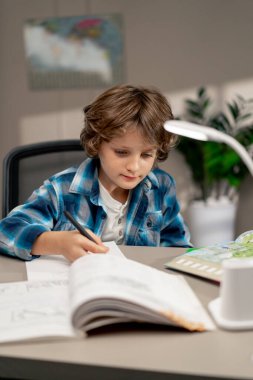 Çocuk odasında beyaz kazaklı ve mavi kareli tişörtlü bir okul çocuğu ödevlerini bir deftere yazarak yapıyor.