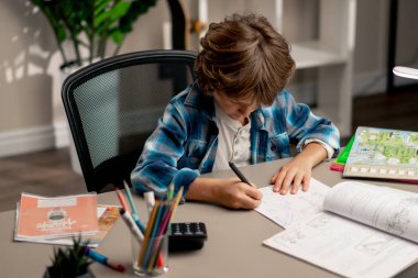 Çocuk odasında beyaz kazaklı ve mavi kareli tişörtlü bir okul çocuğu ders çalışmalarını not defterine yazarak yapıyor.