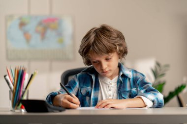 Çocuk odasında beyaz kazaklı ve mavi kareli tişörtlü bir okul çocuğu ödevlerini bir deftere yazarak yapıyor.