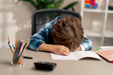 Çocuk odasında beyaz kazaklı ve mavi kareli tişörtlü bir çocuk başının arkasındaki masaya uzanmış. Sıkıntılı duygular çeken çocuklar için zor bir görev.