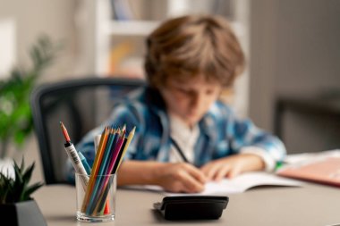 Çocuk odasında beyaz kazaklı ve mavi kareli tişörtlü bir okul çocuğu ödevlerini bir deftere yazarak yapıyor.