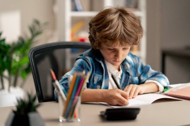 Çocuk odasında beyaz kazaklı ve mavi kareli tişörtlü bir okul çocuğu ödevlerini bir deftere yazarak yapıyor.