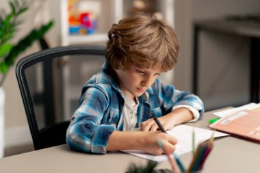 Çocuk odasında beyaz kazaklı ve mavi kareli tişörtlü bir okul çocuğu ödevlerini bir deftere yazarak yapıyor.