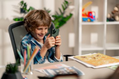 Çocuk odasında beyaz kazaklı ve mavi kareli gömlekli bir okul çocuğu ödevlerini yapmak yerine telefonda oturuyor.