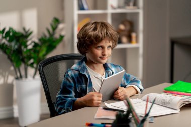 Çocuk odasında beyaz kazaklı ve mavi kareli tişörtlü bir öğrenci tablet modern eğitimin ödevlerini okuyor.
