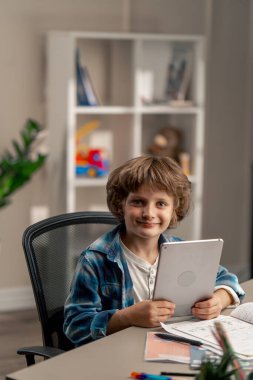 Çocuk odasında beyaz kazaklı ve mavi kareli tişörtlü bir okul çocuğu fotoğraf makinesine bakarak tablet modern eğitimin ödevlerini okuyor.